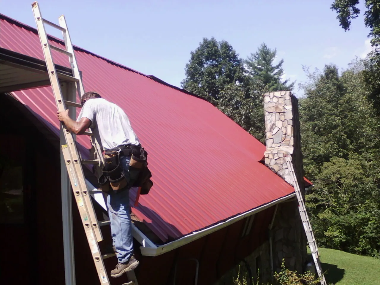 Roofer on a ladder beside a red metal roof and stone chimney — Soffit & Fascia Repair by Roofing Brothers in Ivins